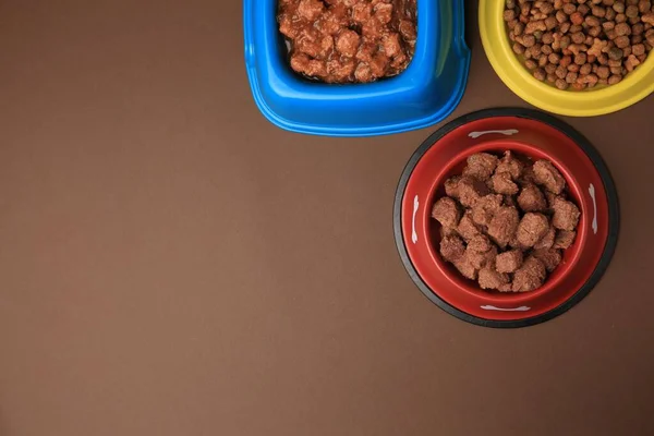 Dry and wet pet food in feeding bowls on brown background, flat lay. Space for text