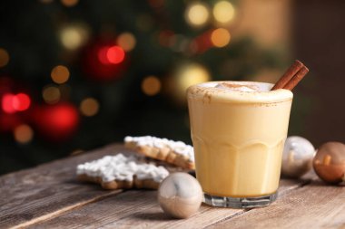 Tasty eggnog with cinnamon, cookies and baubles on wooden table against blurred festive lights. Space for text