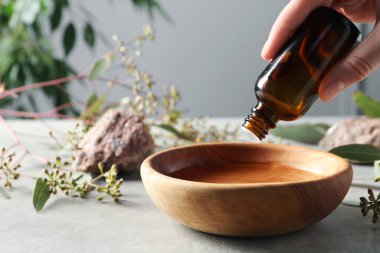 Kadın açık gri masada şişeden kaseye okaliptüs yağı damlatıyor, yakın plan. Metin için boşluk