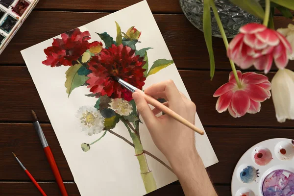 Woman painting flowers with watercolor at wooden table, top view. Creative artwork
