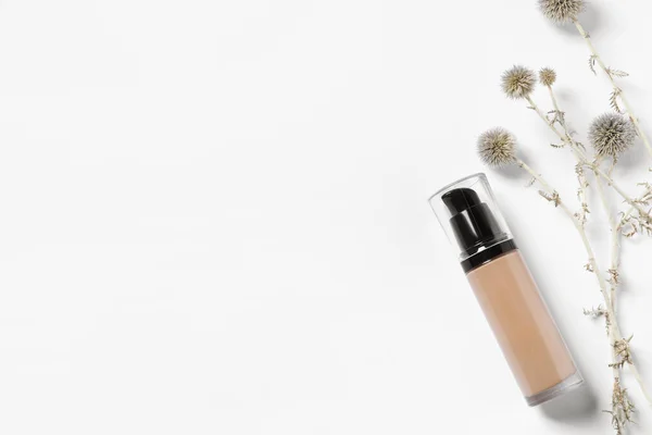 Bottle of skin foundation and decorative flowers on white background, flat lay space for text. Makeup product