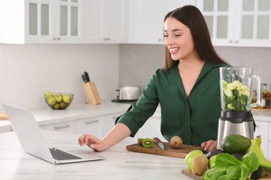 Genç bir kadın mutfaktaki beyaz masada smoothie hazırlarken dizüstü bilgisayar kullanıyor.
