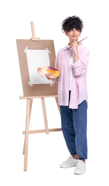 在白色背景的画架附近拿着画笔和艺术家的调色板的年轻女子 — 图库照片