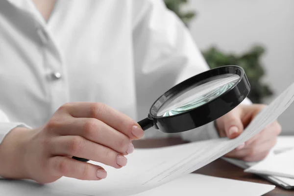 Woman looking at document through magnifier at table indoors, closeup. Searching concept