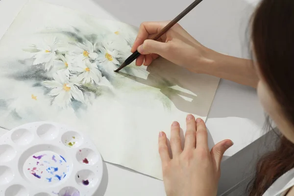 Woman painting flowers with watercolor at white table, above view. Creative artwork