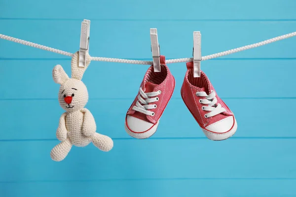 Cute baby sneakers and crochet toy drying on washing line against light blue wooden wall