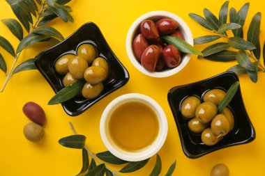 Sarı arka planda bir kase yağ, zeytin ve ağaç dalları, düz uzanmış.