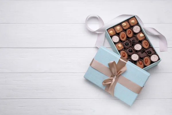Open box of delicious chocolate candies and color ribbon on white wooden table. flat lay. Space for text