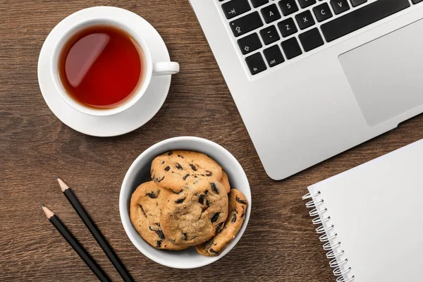 Chocolate chip cookies, cup of tea, office supplies and laptop on wooden table, flat lay