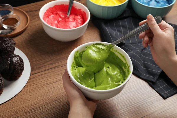 Woman mixing cream with green food coloring at wooden table, closeup
