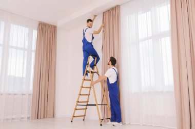 Üniformalı işçiler pencere perdelerini içeri asıyorlar.