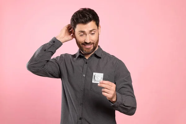 Confused man holding condom on pink background. Safe sex
