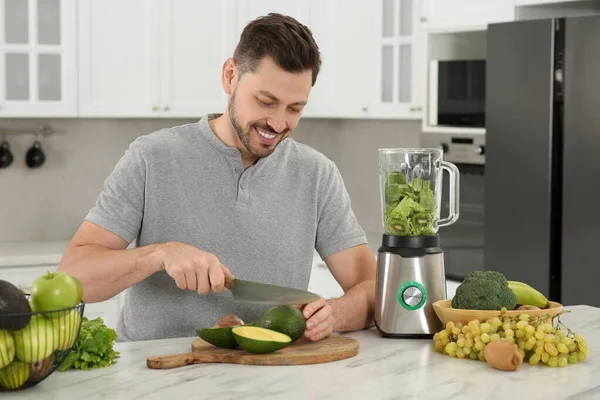 stock image Happy man cutting avocado for delicious smoothie at white marble table in kitchen