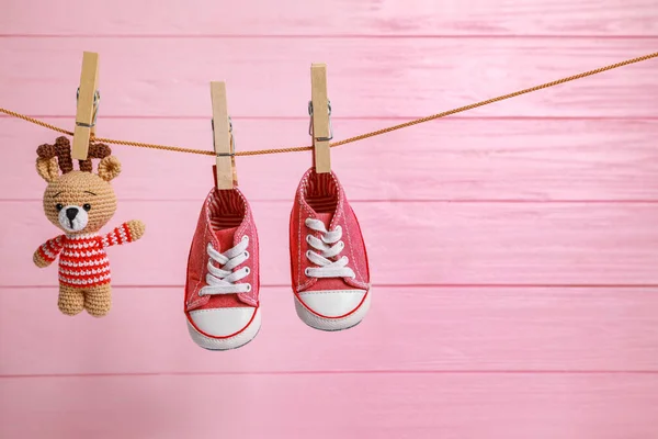 Cute baby sneakers and crochet toy drying on washing line against pink wooden wall. Space for text