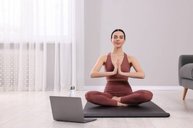 Spor kıyafetli bir kadın evde dizüstü bilgisayarın yanında meditasyon yapıyor, mesaj için yer var.