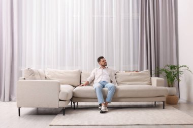 Oturma odasındaki güzel perdelerle pencerenin yanındaki kanepede dinlenen mutlu adam.