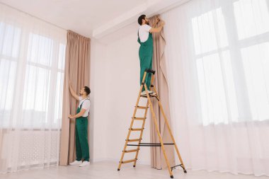 Üniformalı işçiler pencere perdelerini içeri asıyorlar.