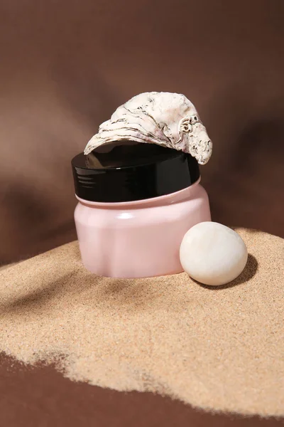 Jar of body cream, seashell and stone on sand against brown background