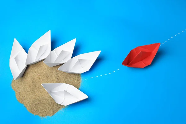 Red paper boat floating away from others on light blue background, flat lay. Uniqueness concept