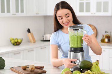 Beautiful young woman preparing tasty smoothie at white table in kitchen