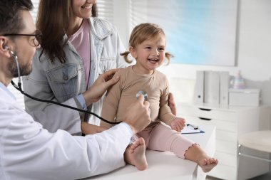 Annesi ve sevimli bebeğinin klinikteki çocuk doktoruyla randevusu var. Doktor küçük kızı muayene ediyor.