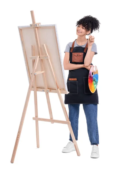 年轻女子在画架附近拿着画笔和艺术家的调色板 画架上有白色背景的帆布 — 图库照片