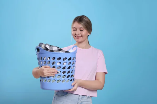 Happy woman with basket full of laundry on light blue background