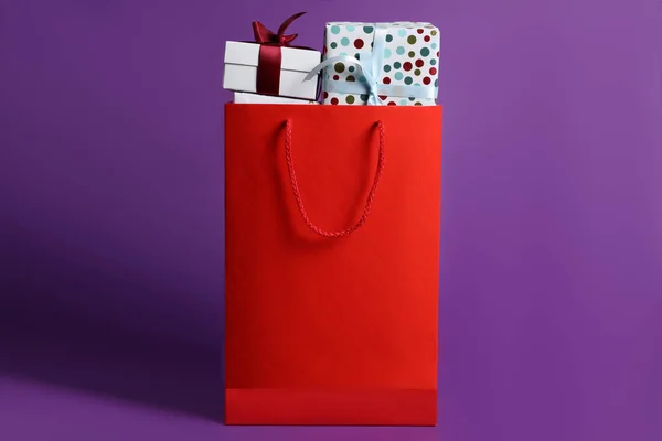 Red paper shopping bag full of gift boxes on purple background