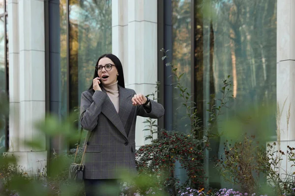 Emotional woman checking time while talking on smartphone outdoors. Being late concept