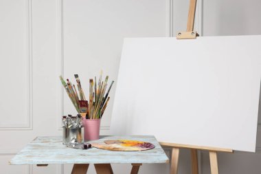 Beyaz duvarın yanındaki ahşap masada boş tuval ve farklı sanat malzemeleriyle.