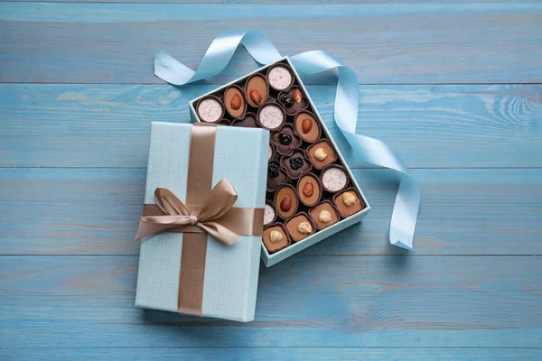 Open box of delicious chocolate candies and color ribbon on light blue wooden table, flat lay