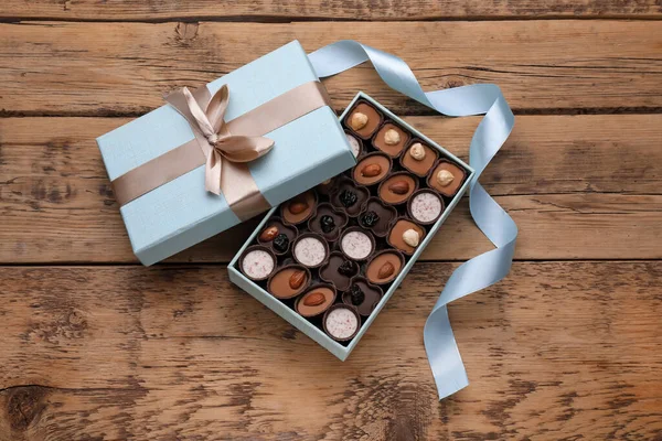 Open box of delicious chocolate candies and light blue ribbon on wooden table, flat lay