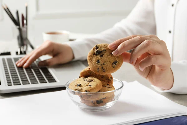 Woman taking chocolate chip cookie from bowl while working with laptop in office, closeup