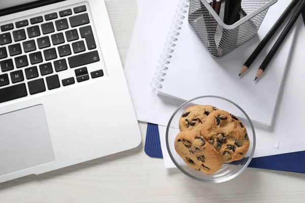 Bowl with chocolate chip cookies, laptop and office supplies on white wooden table, flat lay