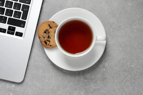 Chocolate chip cookie, cup of tea and laptop on light grey table, flat lay