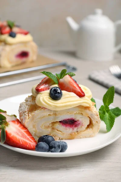 Piece of tasty meringue roll with jam, cream, strawberry, blueberry and mint on white wooden table, closeup