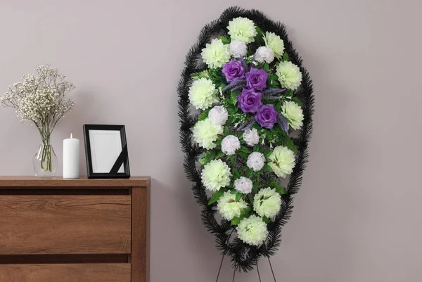 stock image Wreath of plastic flowers and frame with black ribbon, burning candle on commode in room. Funeral attributes