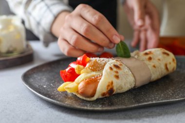 Kadın açık gri masada sosisli, patates kızartması ve biberli nefis pita dürümü pişiriyor.