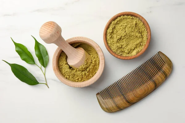 Mortar of henna powder, comb and green leaves on white marble table, flat lay. Natural hair coloring