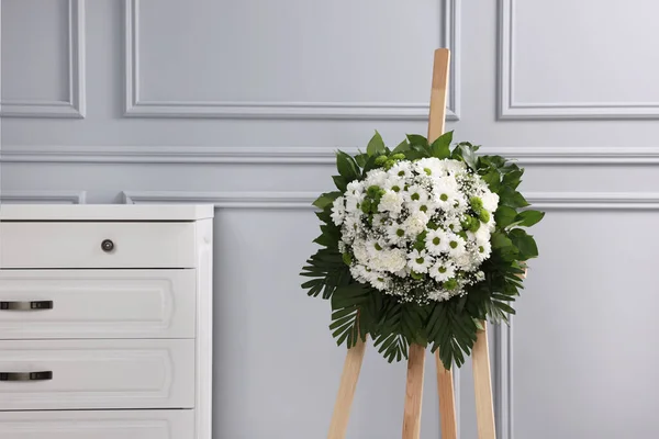 Funeral wreath of flowers on wooden stand near light grey wall indoors
