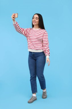 Gülümseyen genç bir kadın açık mavi arka planda akıllı telefonuyla selfie çekiyor.