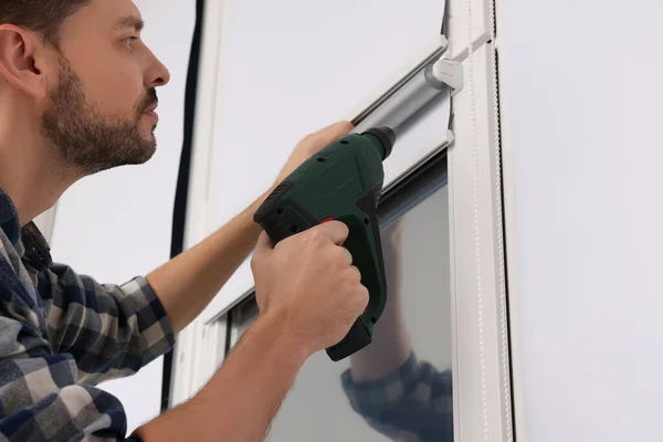 Man with drill installing roller window blind indoors, closeup