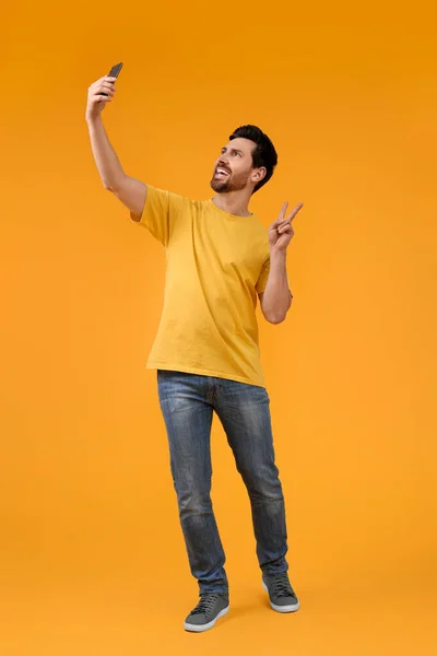 微笑的男人拿着智能手机自私自利 在黄色的背景上展示和平的标志 — 图库照片