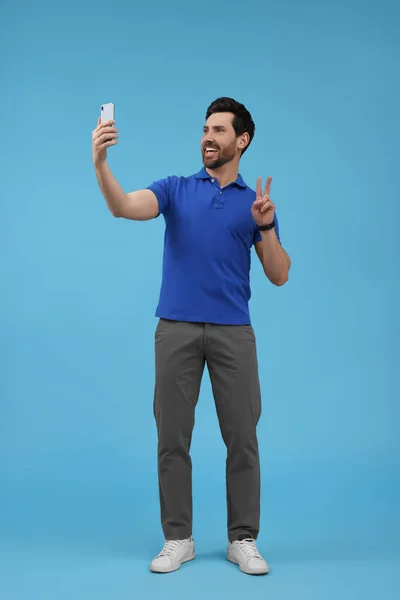Uśmiechnięty Mężczyzna Robi Selfie Smartfonem Pokazuje Znak Pokoju Jasnoniebieskim Tle — Zdjęcie stockowe