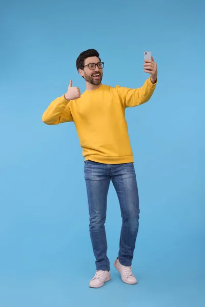 微笑的男人拿着智能手机自拍 用浅蓝色的背景竖起大拇指 — 图库照片