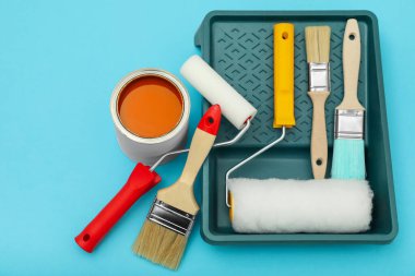 Can of orange paint, brushes, rollers and container on turquoise background, flat lay