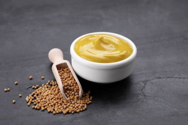Bowl of delicious mustard and scoop with seeds on black textured table clipart