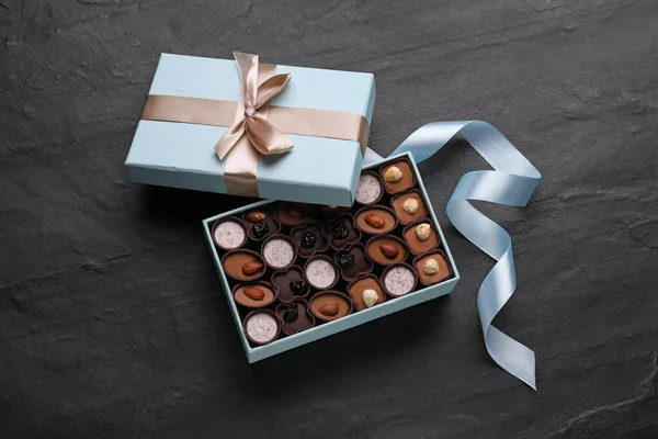 Open box of delicious chocolate candies and light blue ribbon on black table, flat lay
