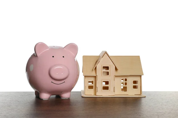 Piggy bank and house model on wooden table against white background. Saving money concept