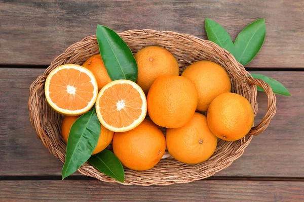 stock image Wicker basket with ripe juicy oranges and green leaves on wooden table, top view